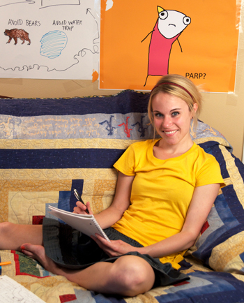 Author Allie Brosh sitting on a bed wearing a yellow t-shirt and a skirt while holding a notebook